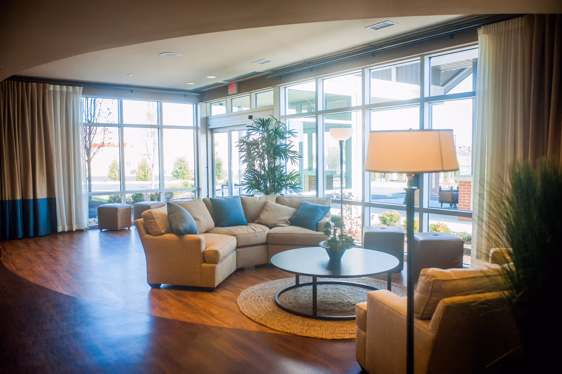 A bright and spacious living room area with large floor-to-ceiling windows letting in natural light. The room features a beige sectional sofa with blue and beige cushions, a round coffee table with a small plant centerpiece, a floor lamp, and additional seating. The floor is wooden, and there are curtains on the windows. Outside the windows, some landscaping and a covered entrance are visible.