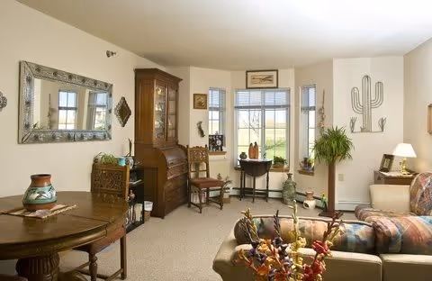 A cozy living room with a beige carpet, a wooden round table with a decorative vase, a large mirror on the wall, a wooden cabinet, a small desk with a chair near a bay window, a couch with patterned cushions, and various decorative items including a large metal cactus wall art and a potted plant.
