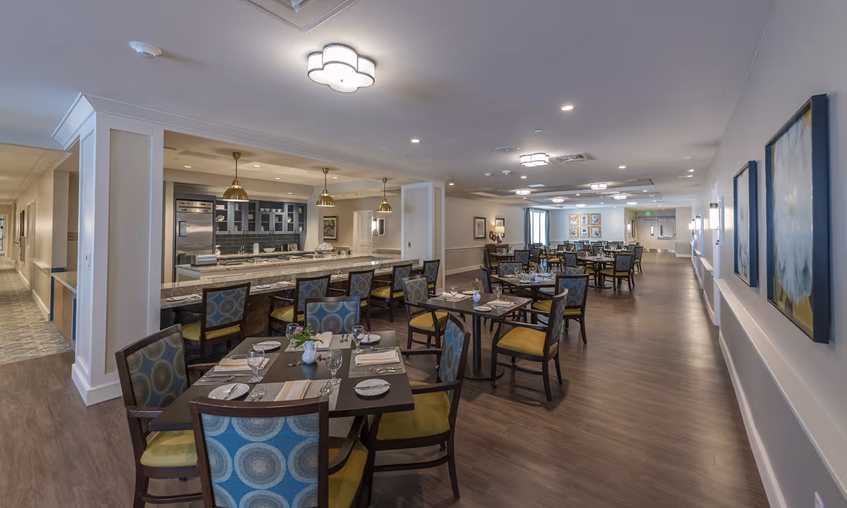 Spacious dining room in Alto Evergreen Walk featuring multiple tables set with plates, glasses, and napkins. Chairs with patterned blue and green upholstery surround the tables. A long counter with bar stools and pendant lights overlooks a kitchen area with stainless steel appliances and cabinetry. The room has wood flooring, soft lighting, and artwork on the walls.