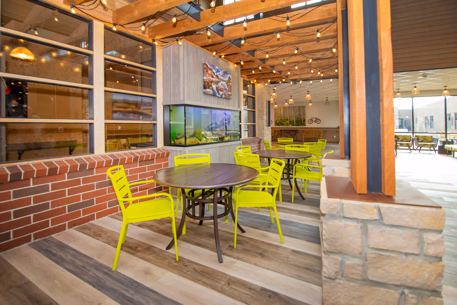 Bright indoor seating area with round tables, neon yellow chairs under wooden beams and string lights, an aquarium and brick half-wall.