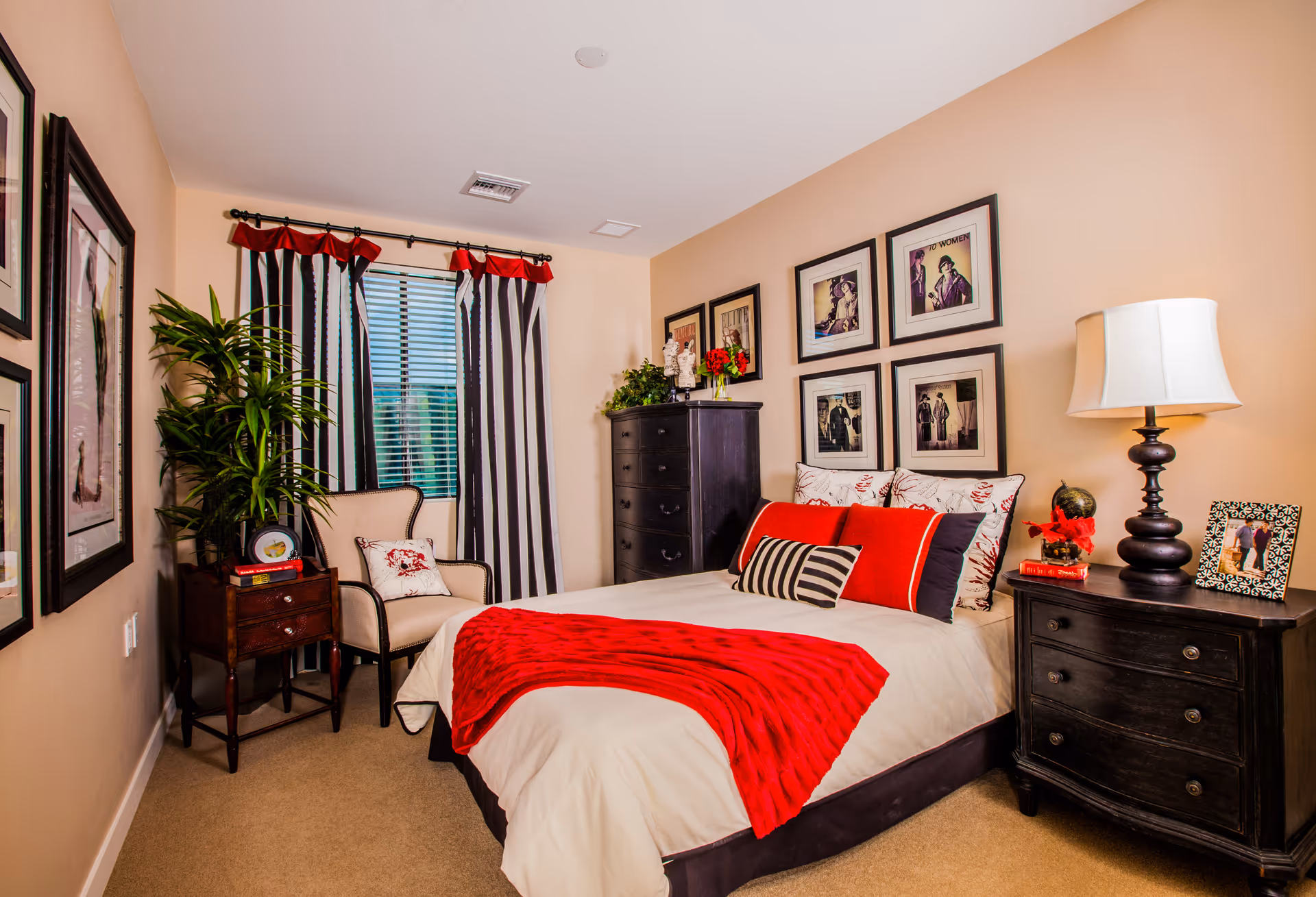 Well-decorated bedroom with a bed dressed in white linens and a red throw, black nightstands, framed wall art, a chair, and striped curtains.