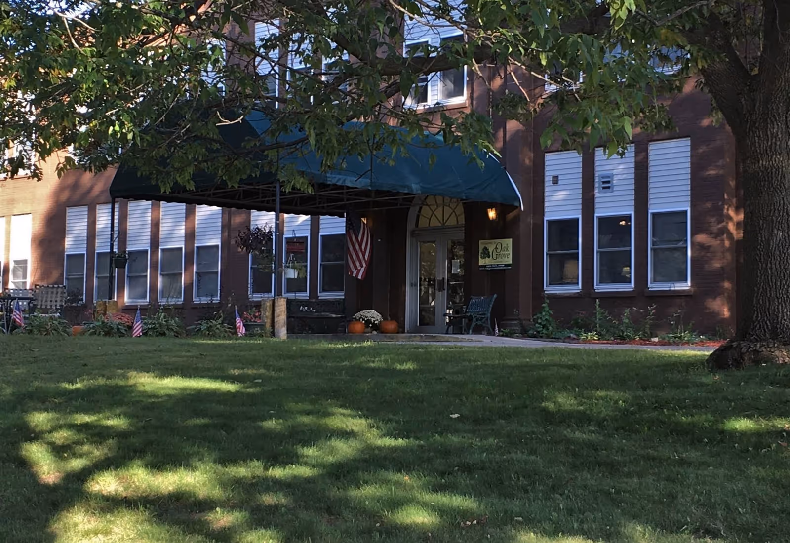 Front entrance of Oak Grove Assisted Care facility with a green canopy above the door, American flag, potted plants, and a large tree casting shadows on the grassy lawn in front.