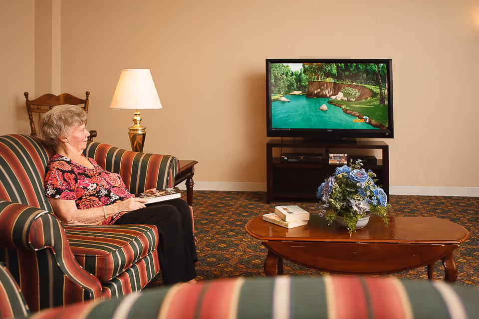 An elderly woman sits on a striped sofa in a cozy living room watching a TV across from a coffee table with flowers and books.