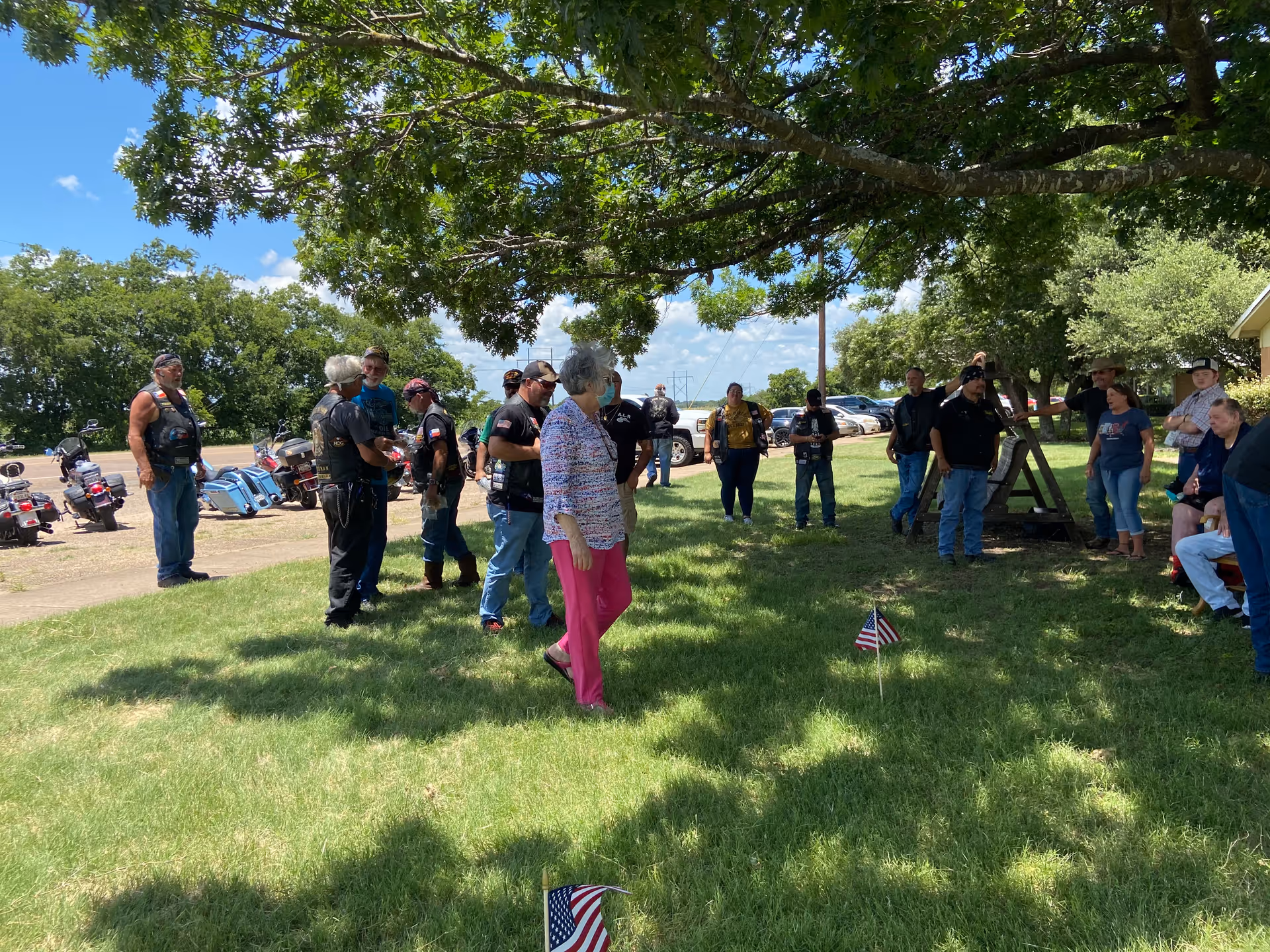 A group of people, including motorcyclists, gathered under a large tree on a sunny lawn with parked motorcycles visible in the background.