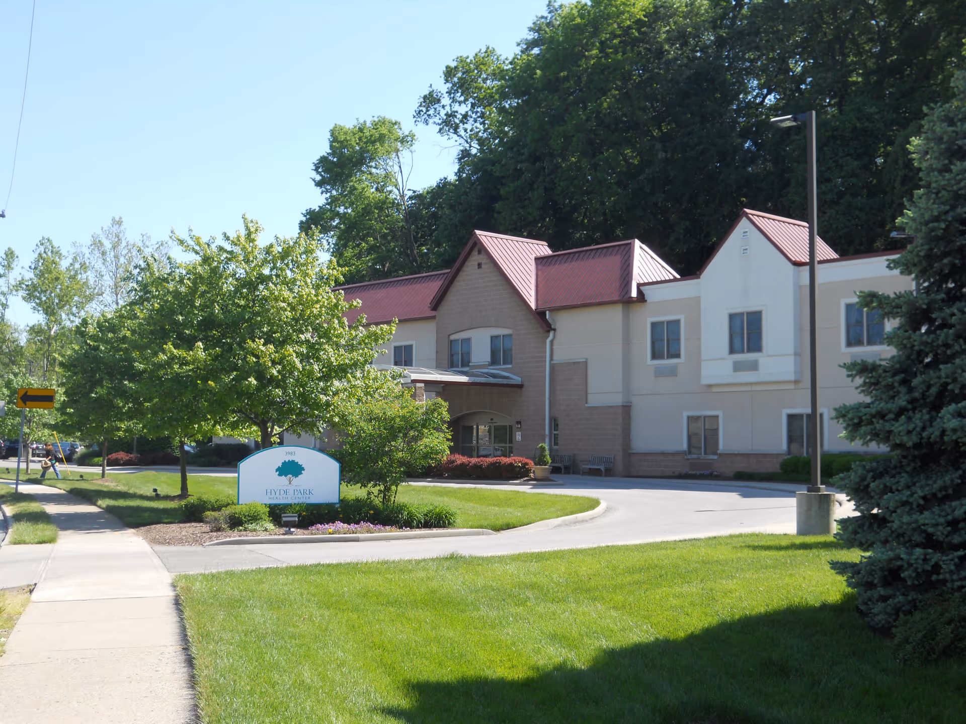 Exterior view of a two-story building with beige and light brown walls and red roofs, surrounded by green trees and a well-maintained lawn under a clear blue sky. A sidewalk and driveway lead to the entrance, with a sign in front reading 'Hyde Park Health Center'.