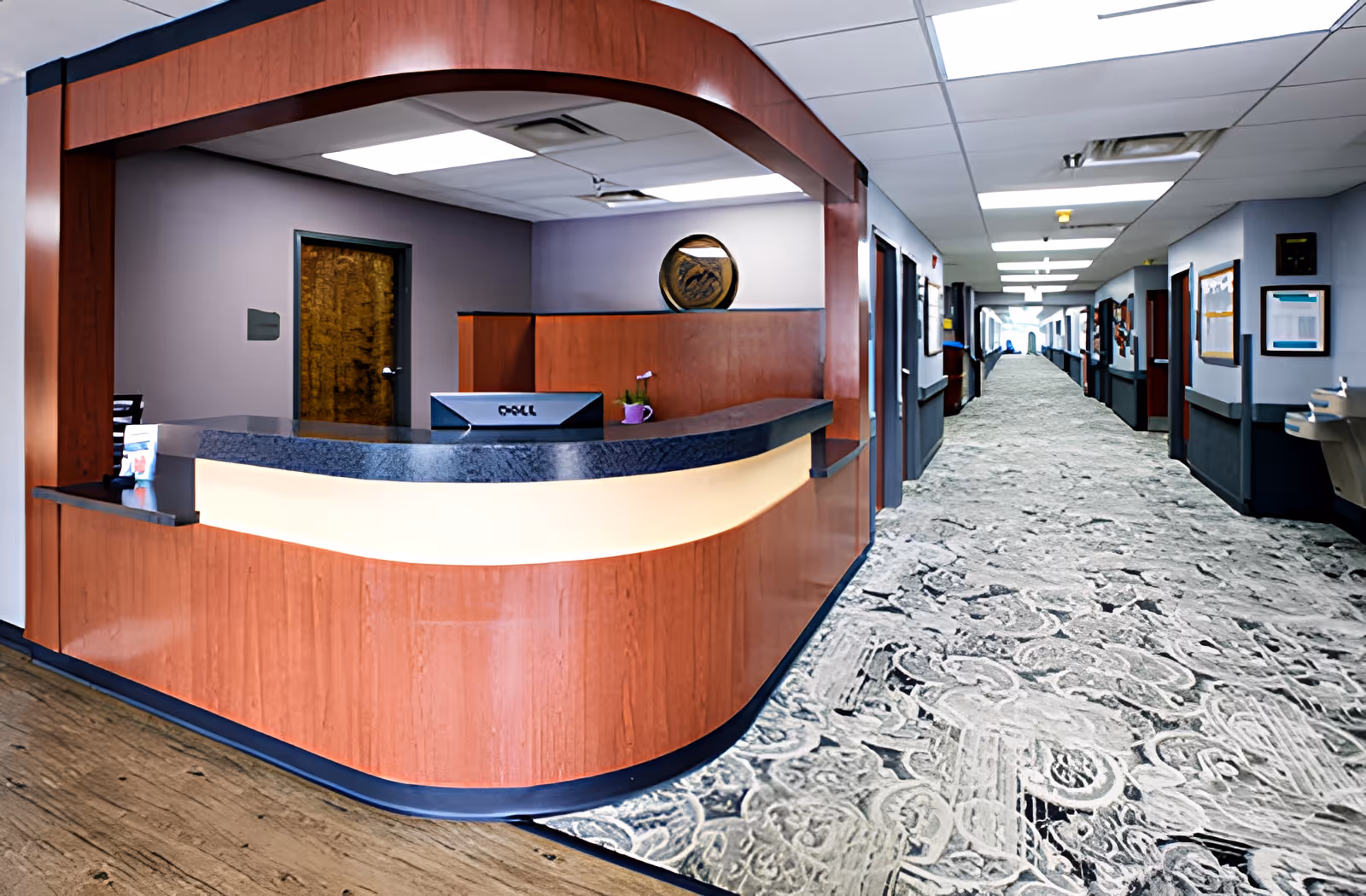 Wood-paneled reception desk with a computer next to a long carpeted hallway inside a senior living facility.