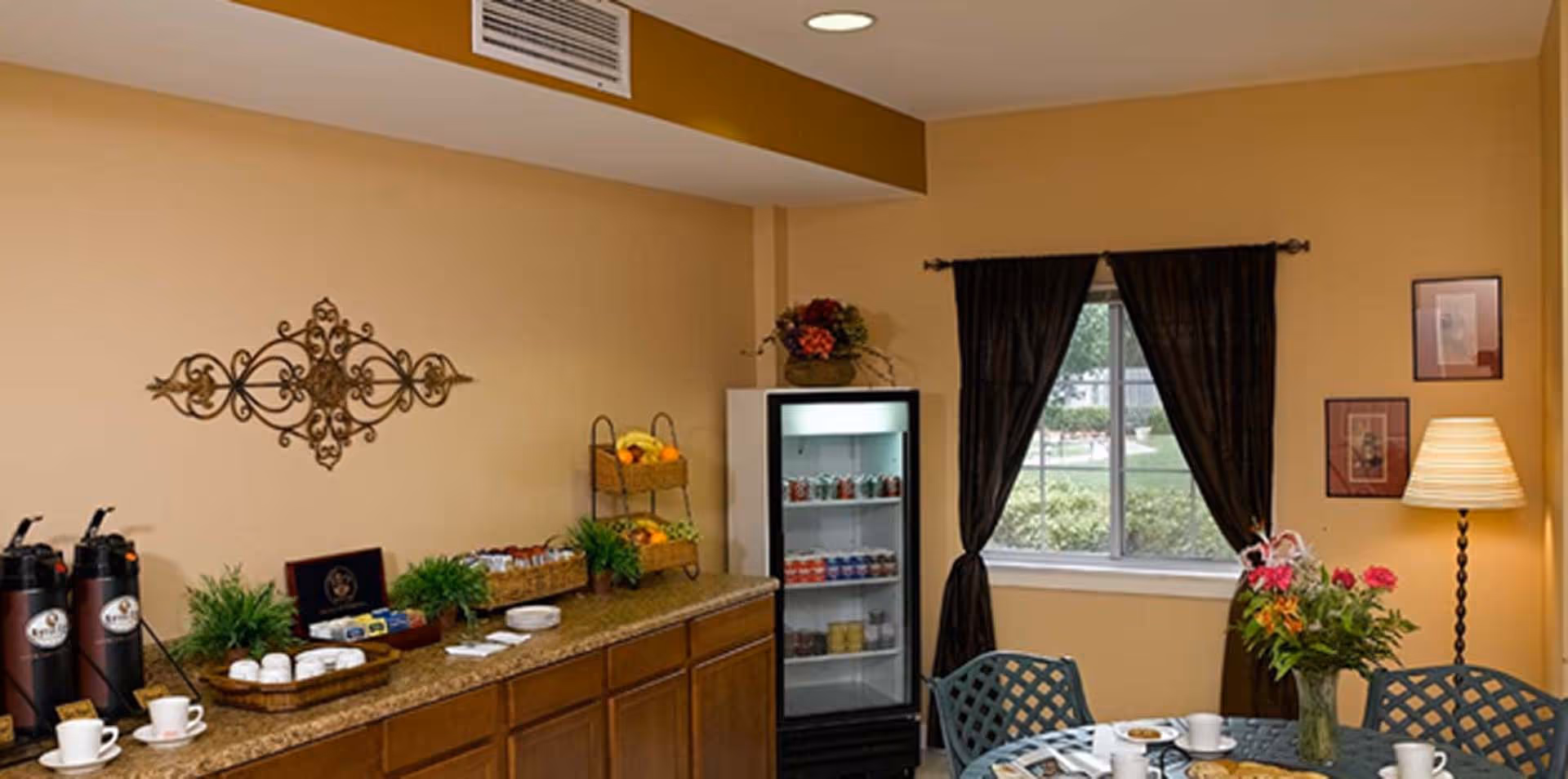 A cozy dining area with a table set for coffee and snacks, featuring a vase with flowers, cups, and plates. To the left, there is a countertop with coffee dispensers, baskets with snacks, and decorative plants. A small refrigerator stocked with drinks is against the wall under a window with dark curtains. The walls are painted beige and decorated with framed pictures and a metal wall ornament.