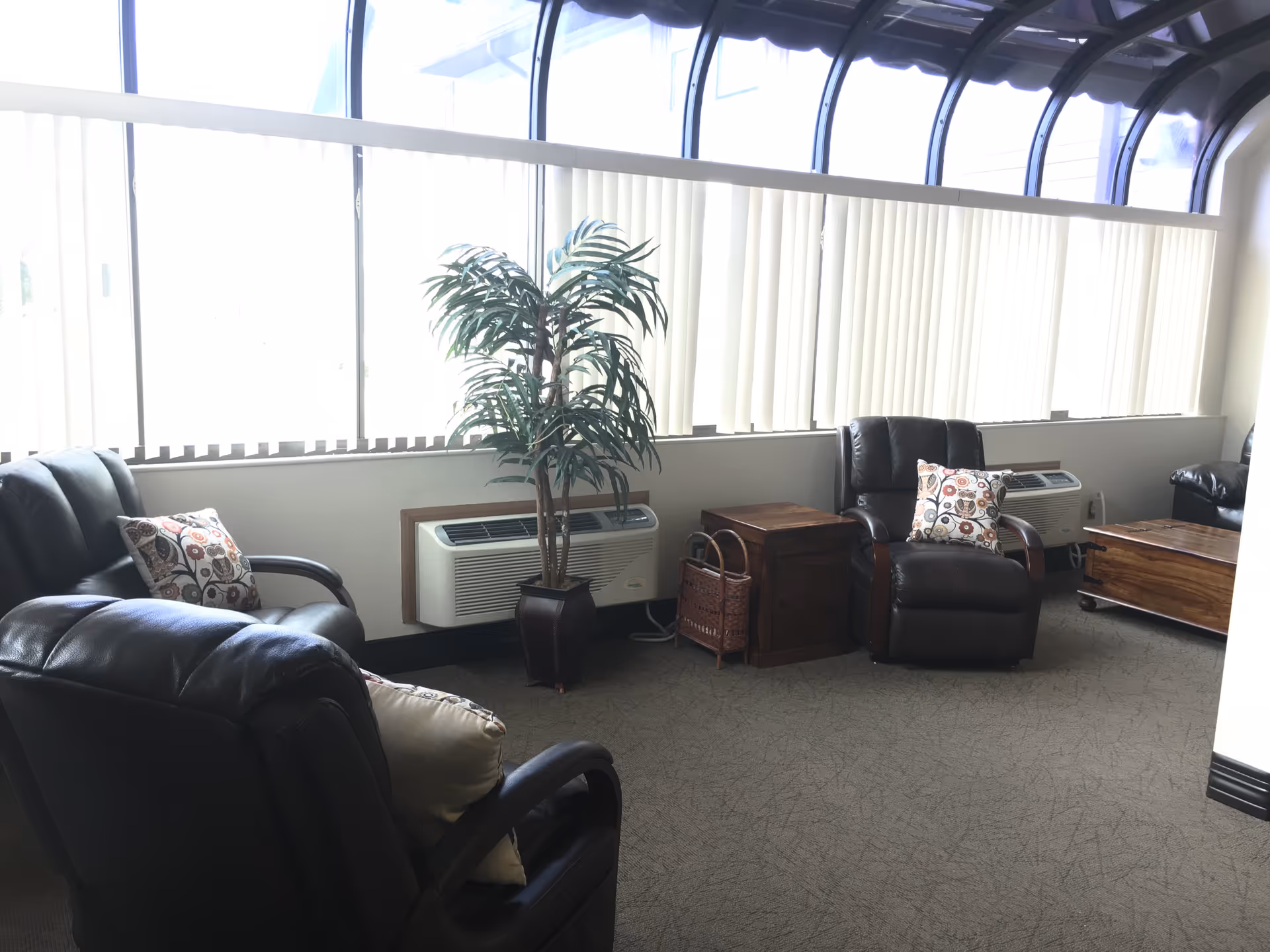 A bright sitting area with large windows covered by vertical blinds, featuring several dark leather armchairs with patterned cushions, a potted plant, wooden side tables, and a wooden chest on wheels.