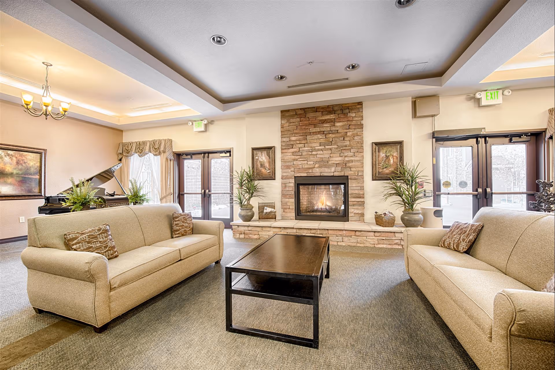 A cozy living room area in an assisted living facility featuring two beige sofas with patterned cushions facing each other across a dark wooden coffee table. A stone fireplace with a fire burning is centered on the far wall, flanked by framed artwork and potted plants. Large windows and glass doors allow natural light to fill the room, and a grand piano is visible in the background.