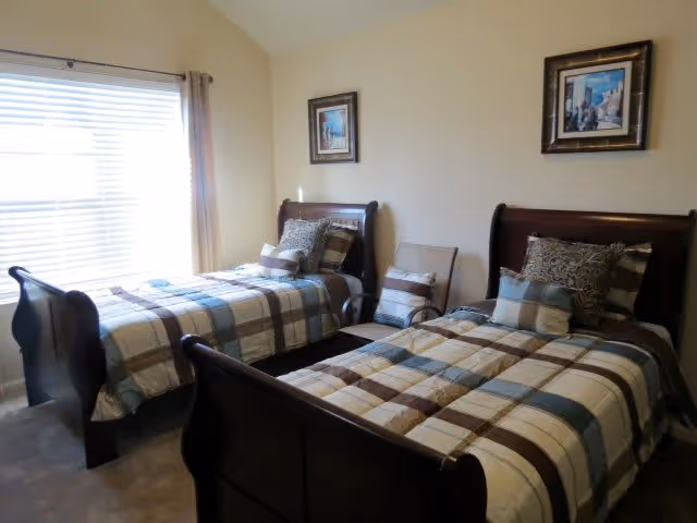 A bedroom with two twin beds featuring dark wooden frames and plaid bedding in shades of blue, brown, and beige. Each bed has multiple pillows. There are two framed pictures hanging on the beige wall above the beds. A window with blinds and beige curtains is on the left side of the room.