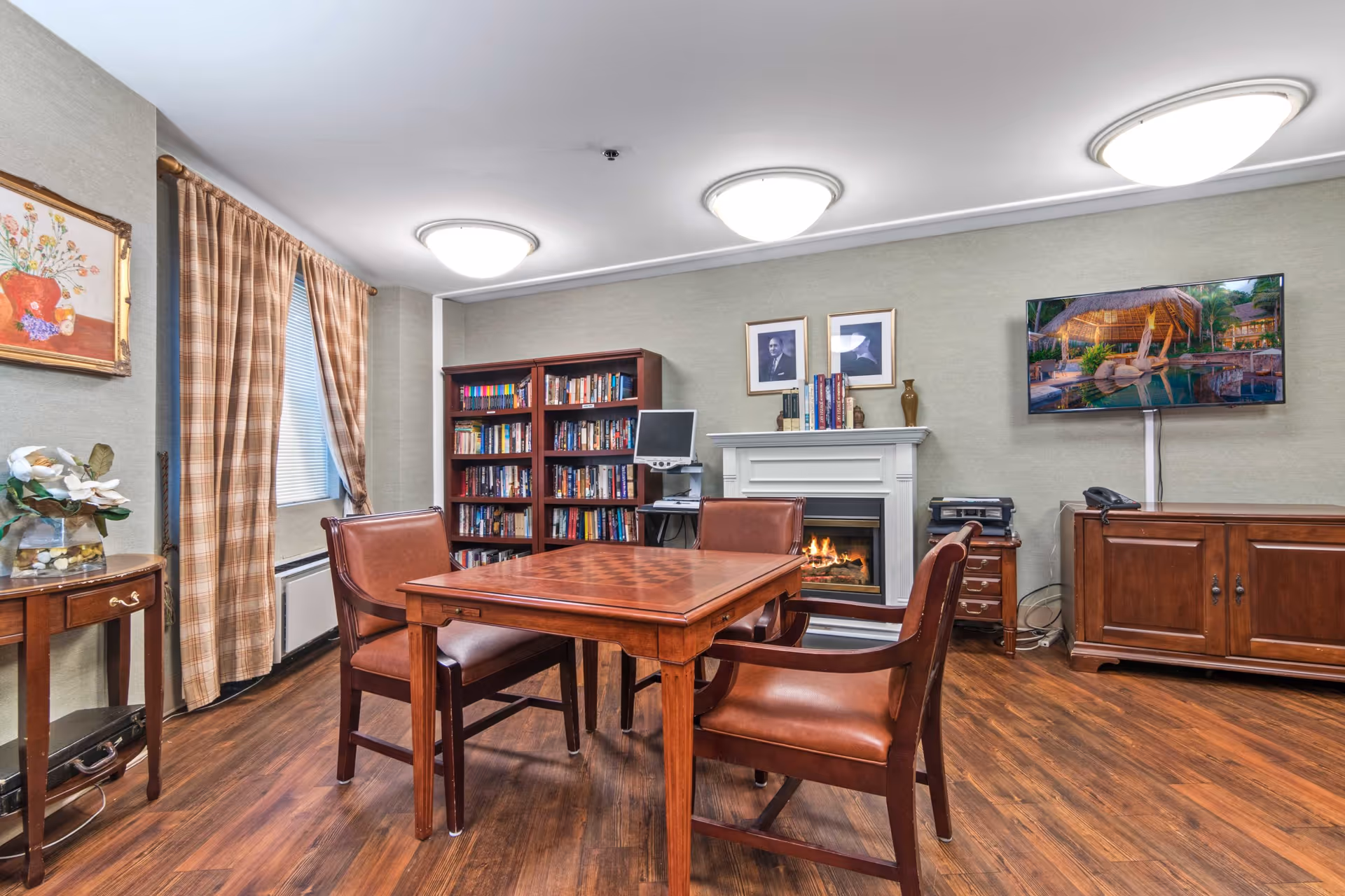 Cozy common room with a wooden table and chairs, a fireplace, bookshelf, and wall-mounted TV.