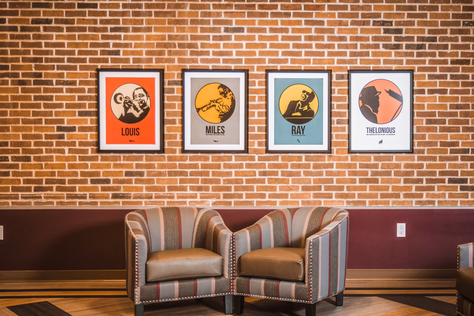 Two striped armchairs placed side by side in front of a brick wall with four framed posters of jazz musicians named Louis, Miles, Ray, and Thelonious.
