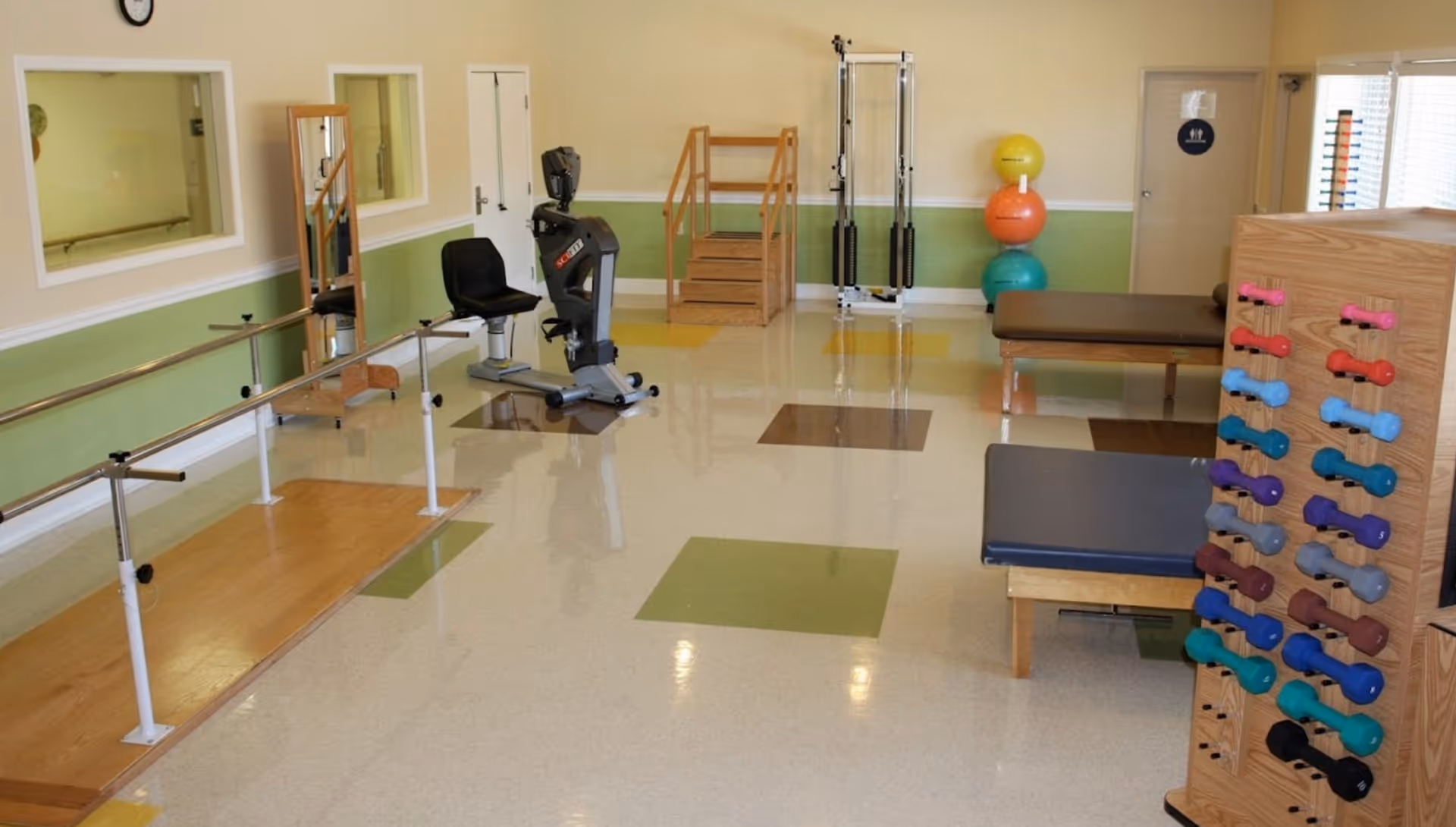 Physical therapy room with parallel bars, exercise bike, therapy stairs, exercise balls, weight rack with colorful dumbbells, and padded therapy tables.