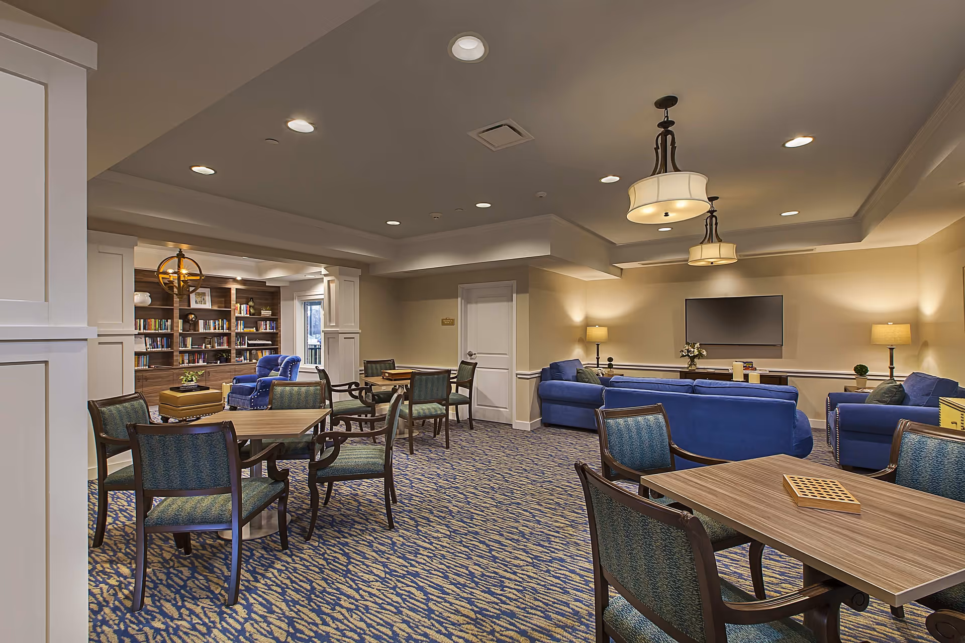 A spacious common area in a senior living facility featuring multiple wooden tables with green cushioned chairs, blue upholstered sofas, a wall-mounted flat-screen TV, lamps, and a bookshelf filled with books. The room has a patterned carpet and recessed lighting with two hanging light fixtures.