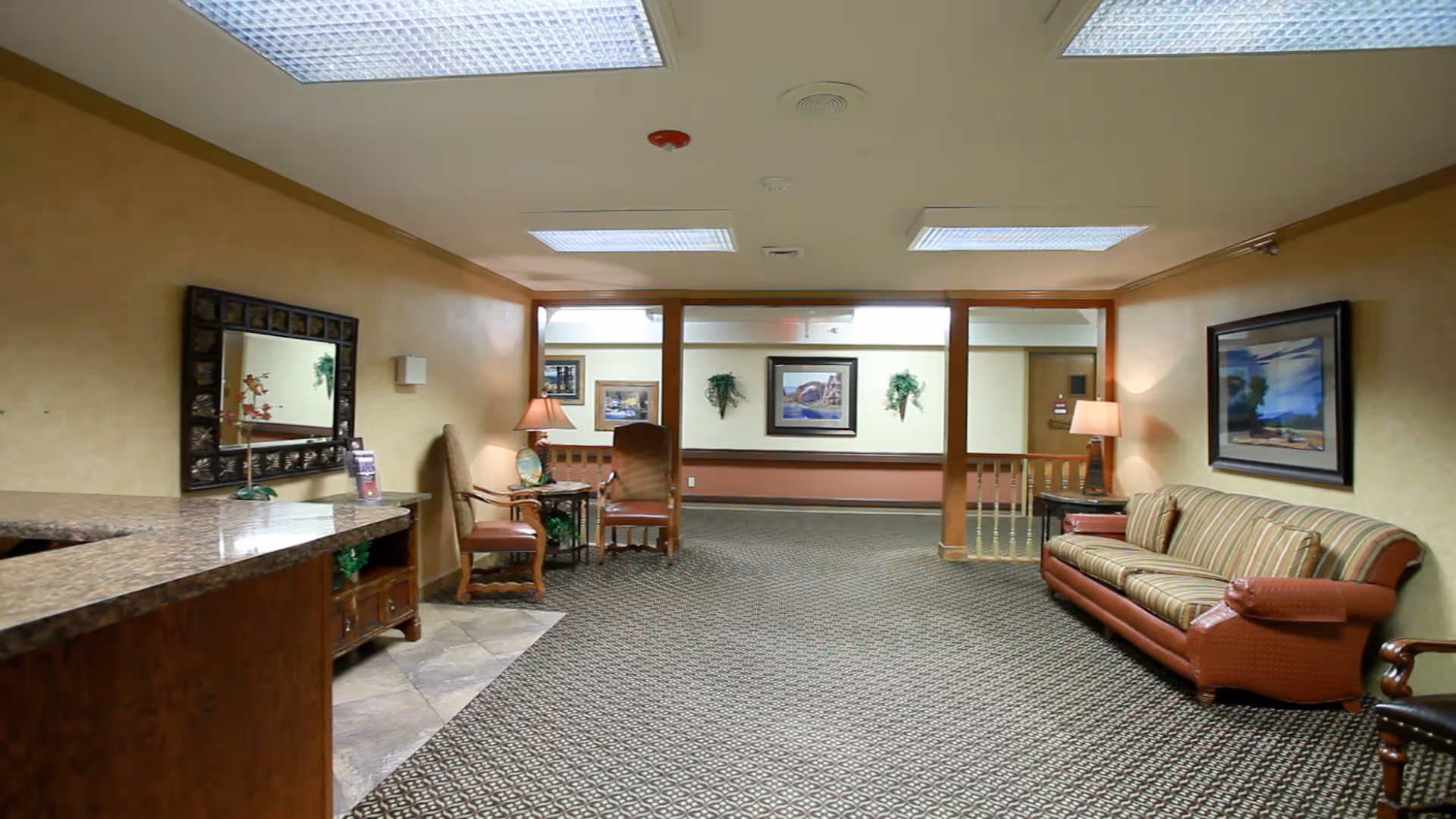 A spacious interior common area with patterned carpet, beige walls, and ceiling lights. The room features a striped sofa on the right, two wooden chairs with cushions near a small table with a lamp, and framed artwork on the walls. A large mirror hangs on the left wall above a wooden counter with a granite top.