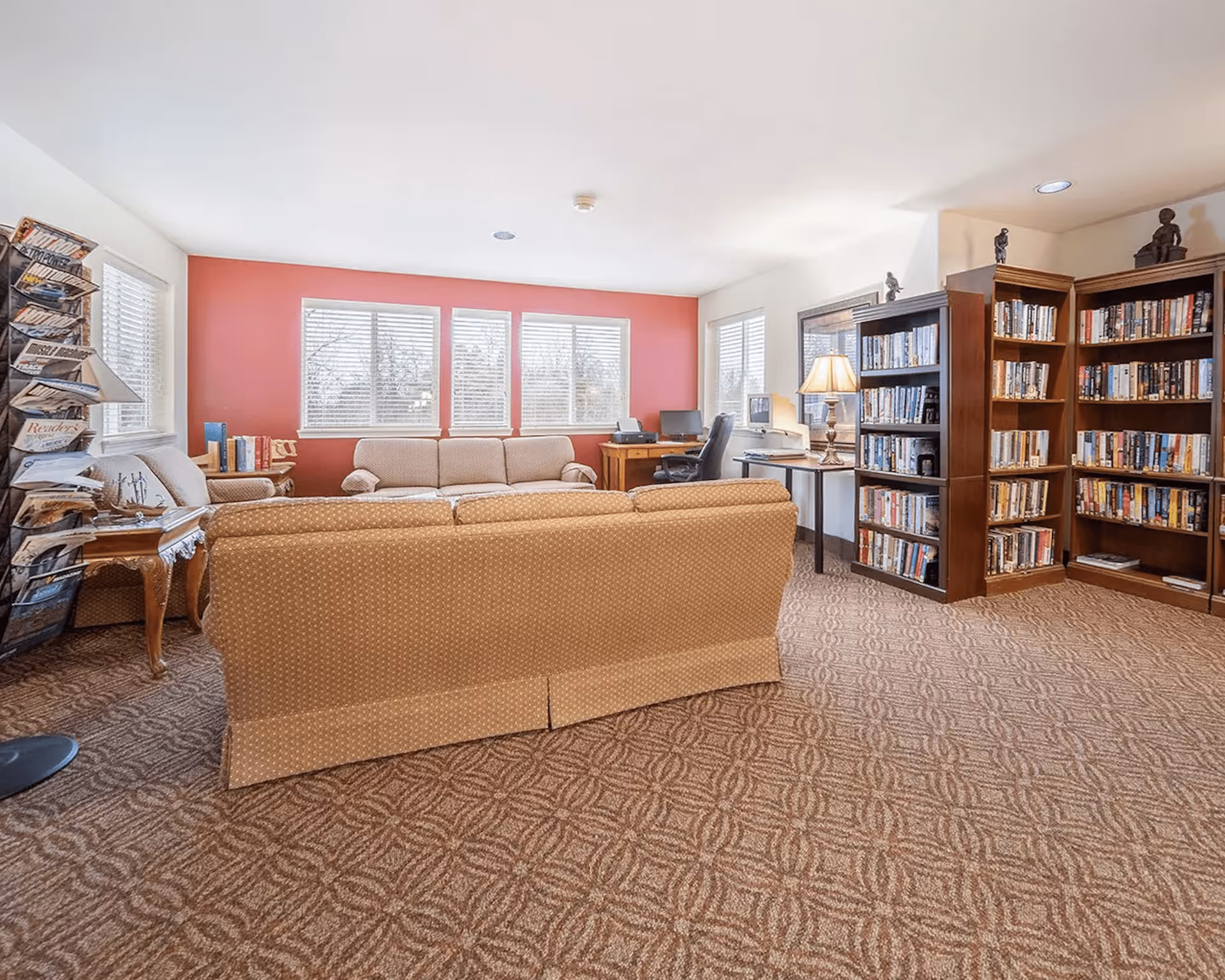 A cozy living room area with patterned carpet, two beige sofas, a wooden side table with a lamp, a magazine rack, and large windows with white blinds letting in natural light. There are bookshelves filled with books and a desk with a computer and office chair near the windows.