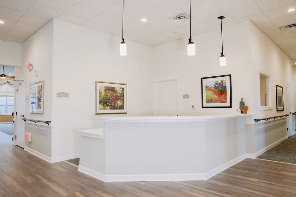 A bright and clean reception area in a senior living facility with a white front desk, three hanging pendant lights, framed artwork on the walls, wood flooring, and handrails along the walls in the hallway.