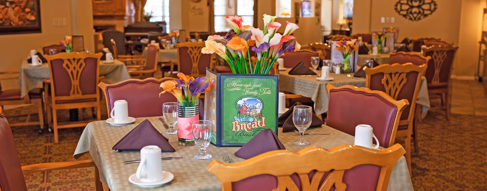 A warmly lit dining room with tables set for meals, featuring floral centerpieces, folded brown napkins, glassware, and white coffee mugs. The tables have beige tablecloths and wooden chairs with red cushioned seats and backs. A menu titled 'The Bread Basket' is prominently displayed on the nearest table.