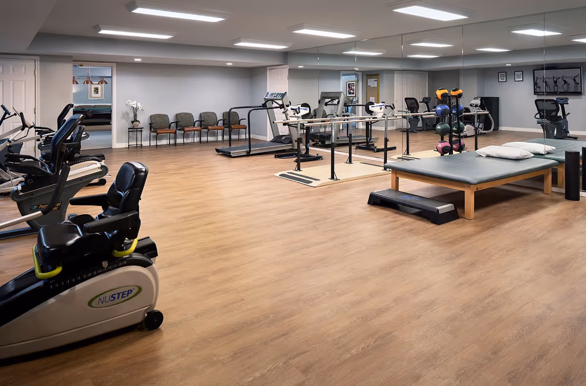 A spacious senior living fitness room with exercise equipment including stationary bikes, treadmills, parallel bars, and a padded exercise table. The room has wood flooring, a large wall mirror, and several chairs lined up against the far wall. The walls are painted light gray and the ceiling has recessed lighting.