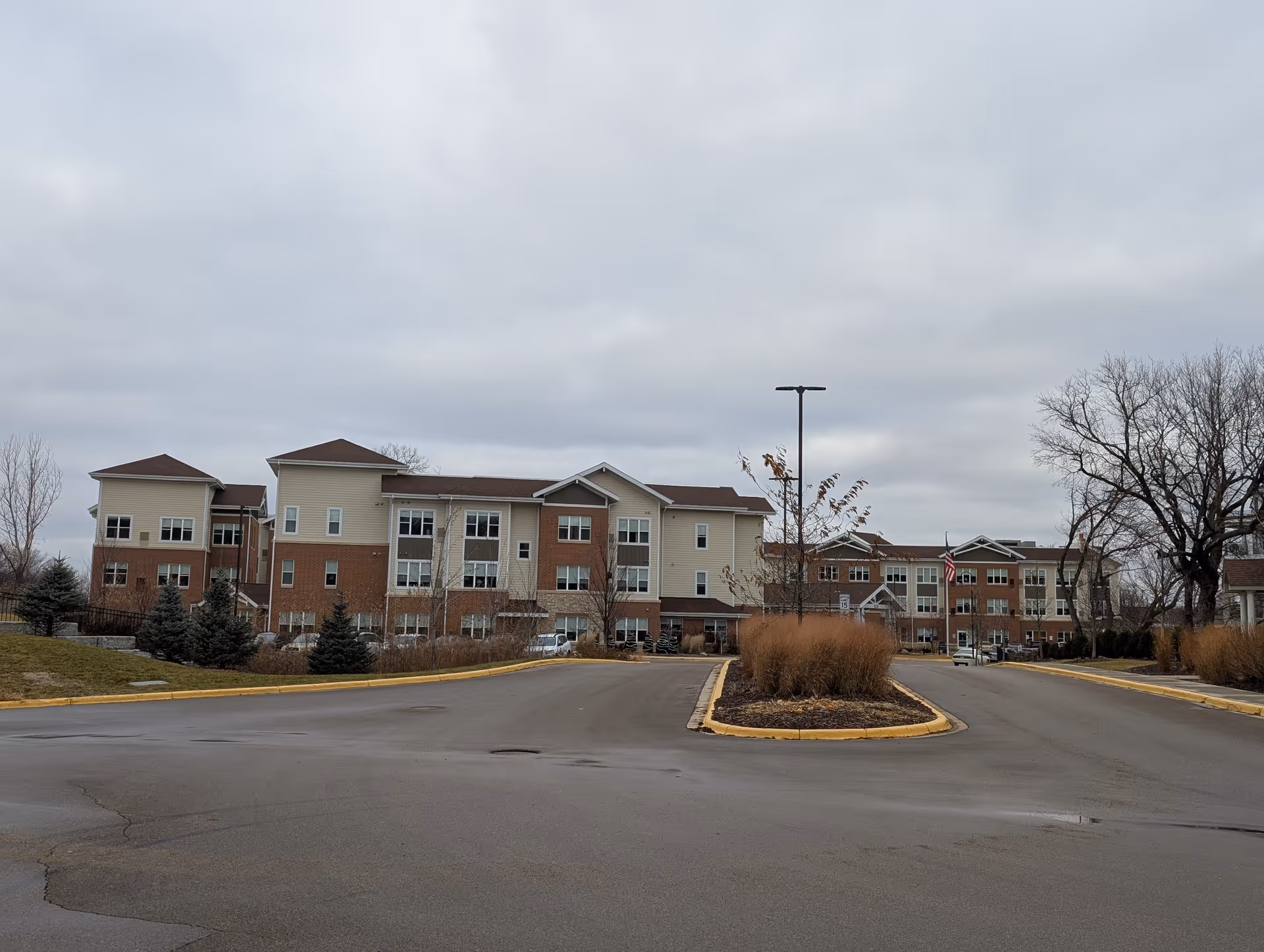 Exterior view of a multi-story senior living facility building with beige and brick facade, surrounded by a parking lot and landscaped areas with trees and shrubs under a cloudy sky.