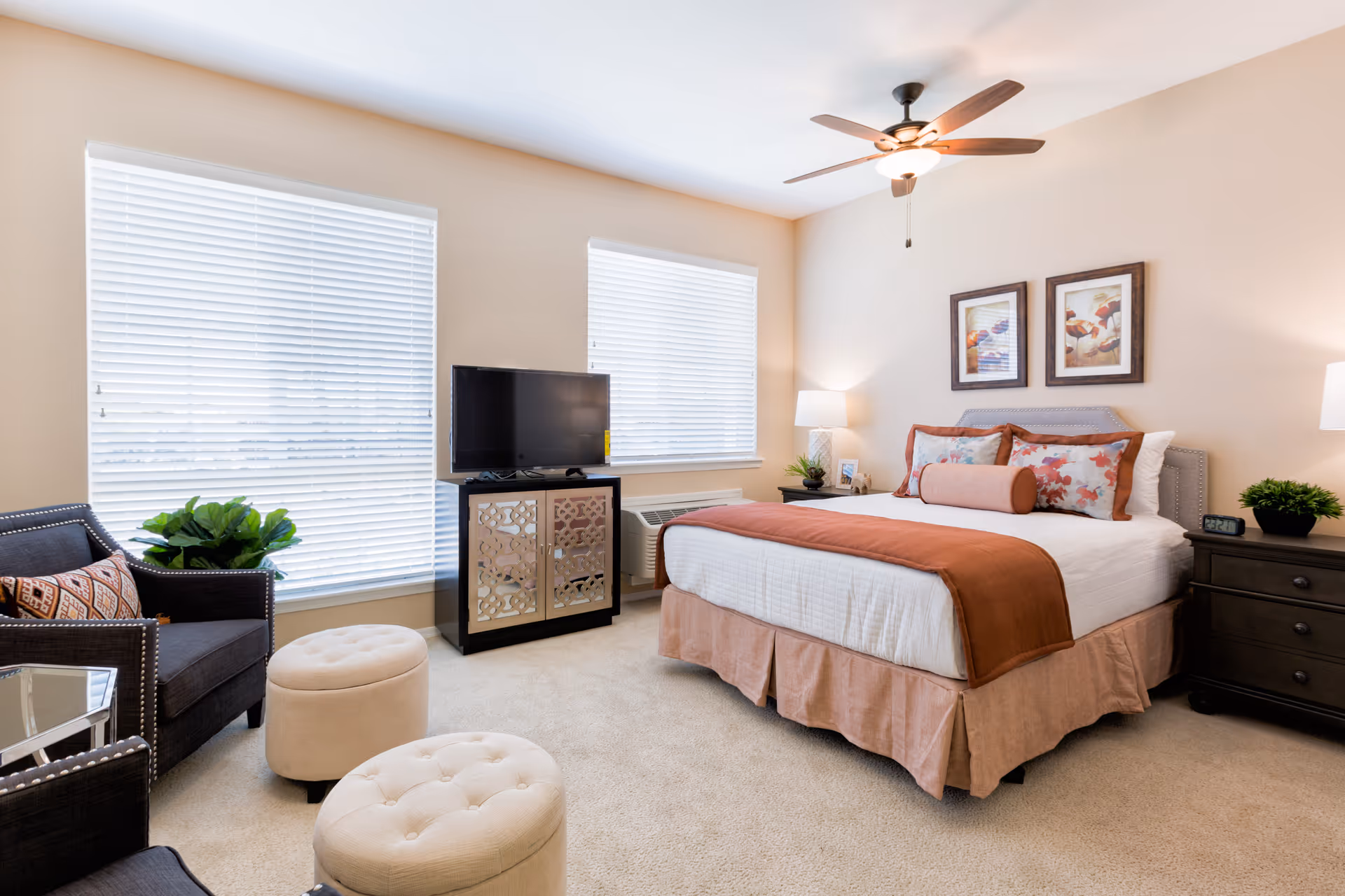 A bright and cozy bedroom with a large bed dressed in white and rust-colored bedding. The room features two windows with white blinds, a ceiling fan with a light, a dark armchair with a patterned pillow, two beige tufted ottomans, a TV on a decorative cabinet, two framed pictures above the bed, and two dark nightstands with lamps and small plants.