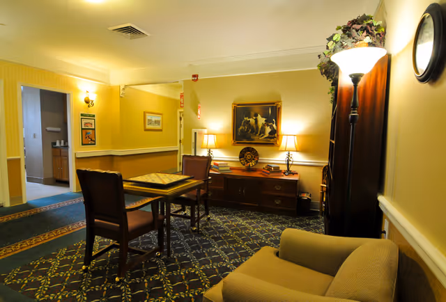 A cozy interior common area with patterned carpet, a small table with two chairs, a cushioned armchair, a wooden cabinet with two table lamps, books, and a decorative plate. There is a painting of dogs on the wall above the cabinet, a tall floor lamp with greenery on top, and a clock on the wall. A doorway leads to another room with a sink and cabinets.