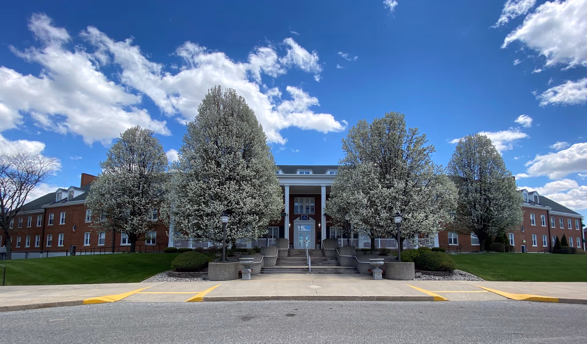 Front view of a red-brick Michigan Masonic Home building with a columned entrance flanked by blossoming trees under a blue sky.