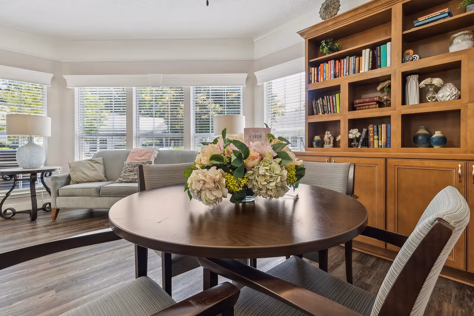 Round wooden dining table with a floral centerpiece in a bright room featuring a sofa, large windows, and built-in wooden bookshelves.