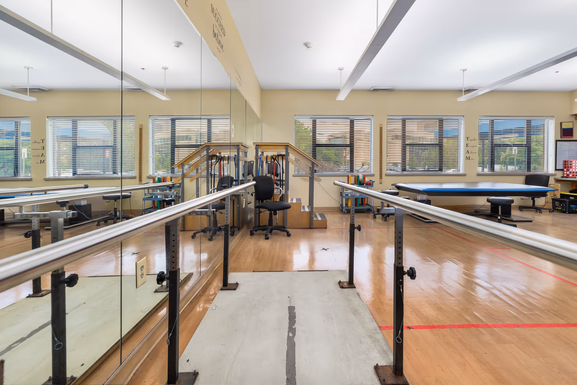 Physical therapy room with parallel bars for walking exercises, a large mirror on one wall, wooden flooring, several windows with blinds, a black office chair, exercise equipment, and a treatment table in the background.
