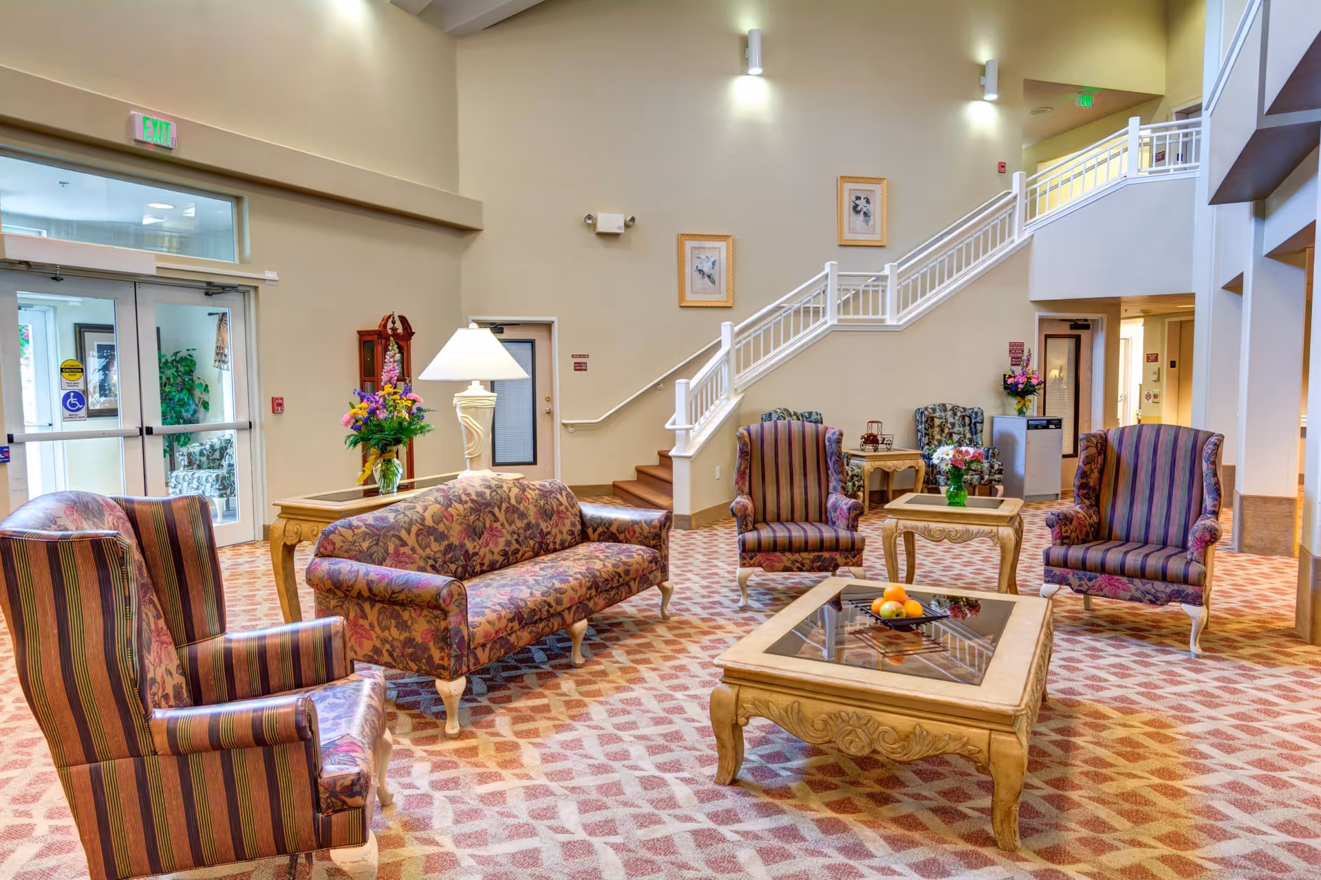 A spacious and well-lit senior living facility common area with patterned carpet, floral and striped upholstered armchairs and sofa, wooden coffee tables with glass tops, a table lamp, and flower arrangements. A staircase with white railings leads to an upper floor, and large glass doors provide natural light from outside.