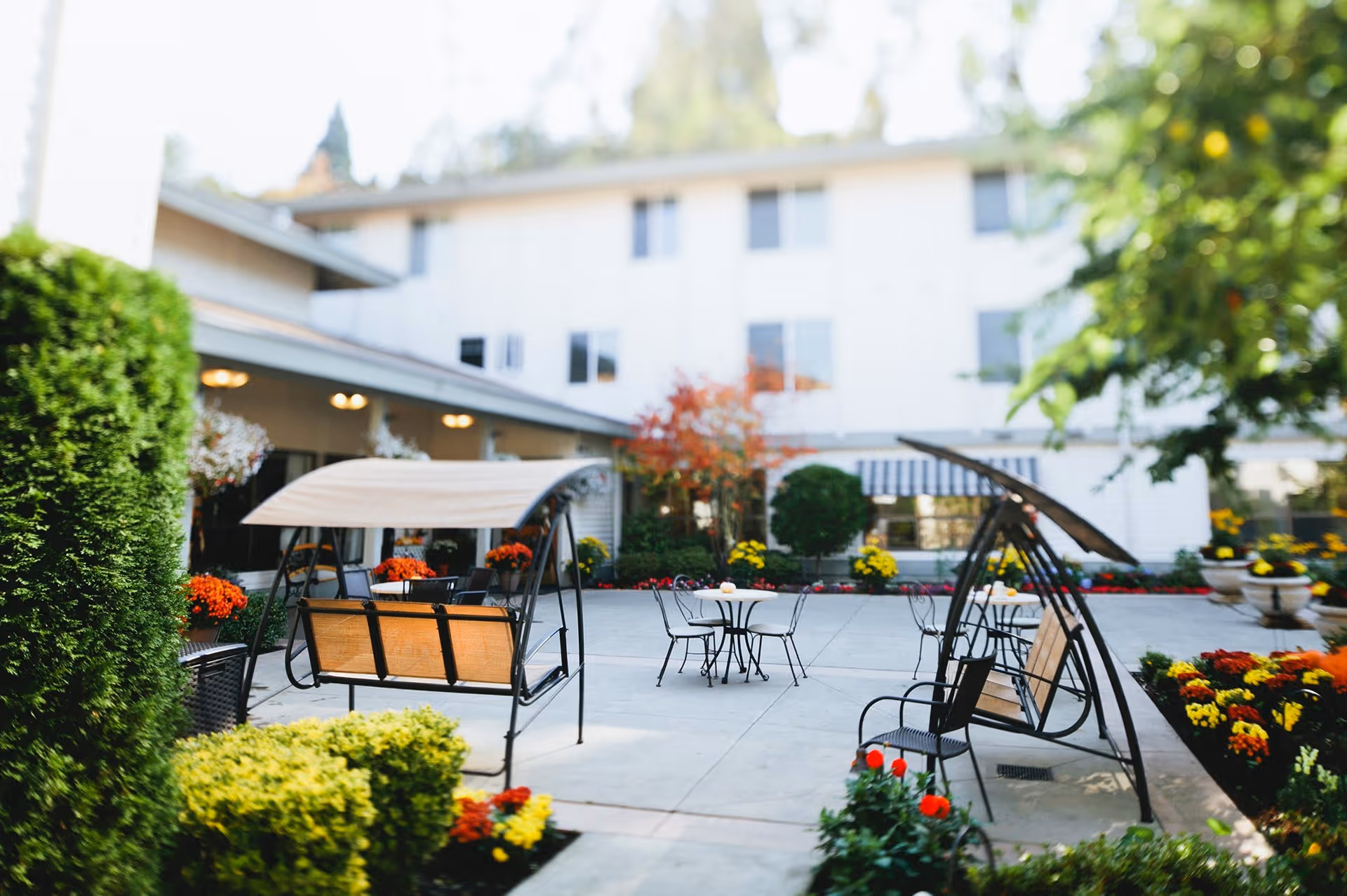 Outdoor patio area at a retirement home with two wooden swing benches with canopies, metal chairs, and small round tables surrounded by colorful flower beds and greenery, with a multi-story building in the background.