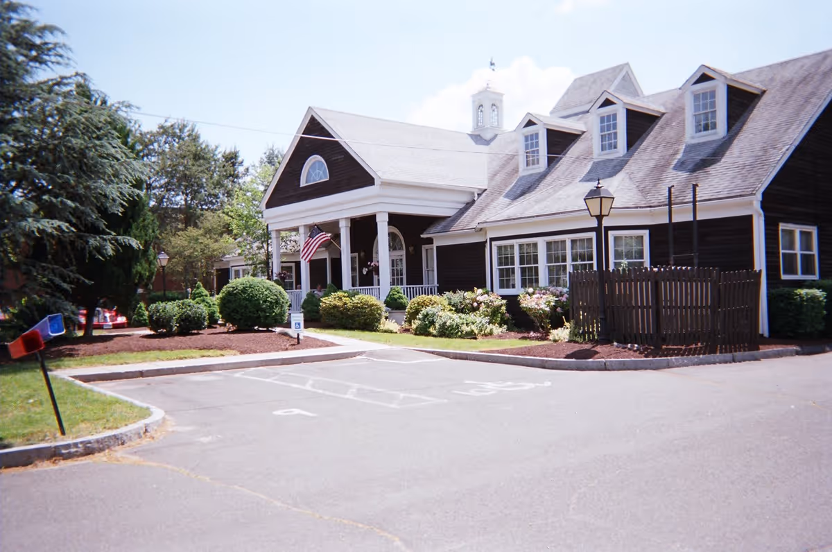 Exterior view of a dark brown building with white trim and a porch, surrounded by landscaped bushes and trees. There is an American flag hanging on the porch, a lamp post near the building, and a parking area with marked spaces in front.