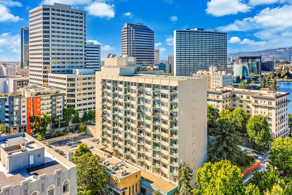 Aerial view of a cityscape featuring a tall residential building with balconies in the foreground, surrounded by other high-rise office buildings and greenery under a blue sky with scattered clouds.