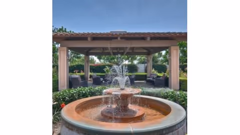 A tiered stone fountain centered in front of a covered outdoor seating pavilion surrounded by plants and trees.