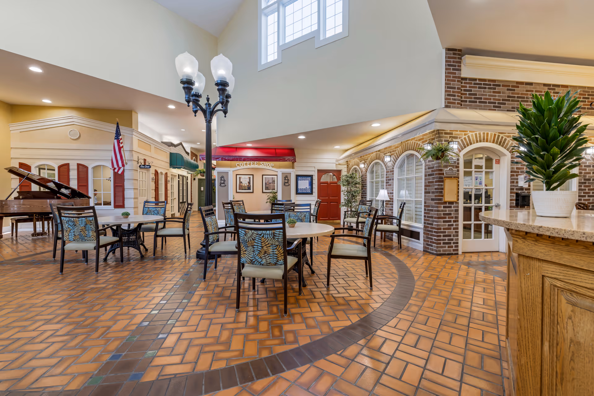 Indoor common area with several round tables and chairs arranged on a patterned tile floor. The space features a high ceiling with large windows, a streetlamp-style light fixture, a grand piano, and storefront-style facades including a coffee shop with a red awning. There are plants and decorative elements, creating a welcoming and cozy atmosphere.