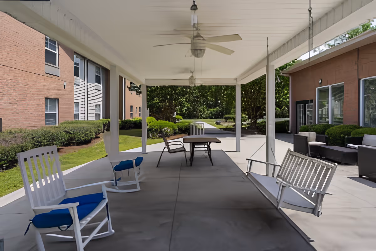 Covered outdoor patio area with white rocking chairs with blue cushions, a white porch swing, a table with chairs, ceiling fans, and greenery surrounding the space. The patio is adjacent to a brick building with windows.
