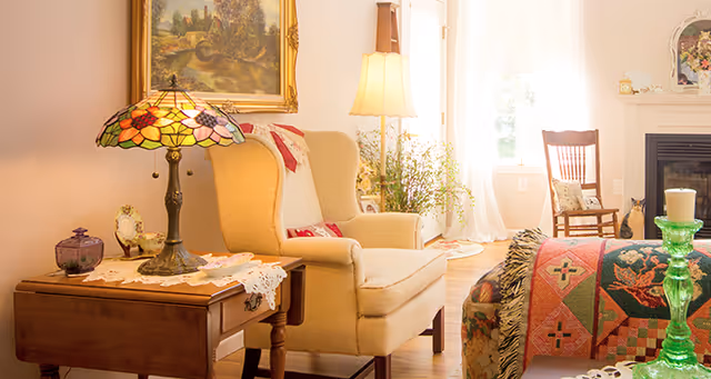 A cozy living room with a beige armchair, a wooden side table with a stained glass lamp, a floor lamp, a wooden rocking chair, and a fireplace. The room is warmly lit with natural light coming through a window with sheer curtains. There is a colorful quilt draped over a piece of furniture and decorative items on the mantel and table.