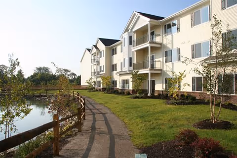 Three-story beige apartment building with balconies beside a pond and a paved walking path.