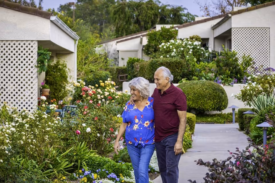 An elderly couple walking arm in arm along a garden pathway surrounded by lush greenery and colorful flowers, with residential buildings in the background.