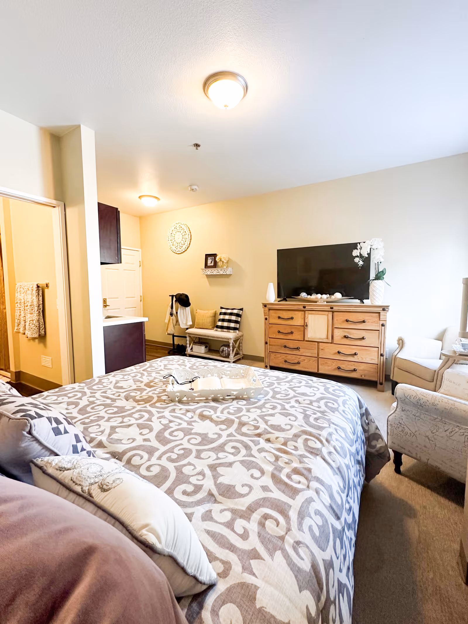 Furnished bedroom with a patterned bedspread, wooden dresser and TV, seating area, and an open doorway to a bathroom.