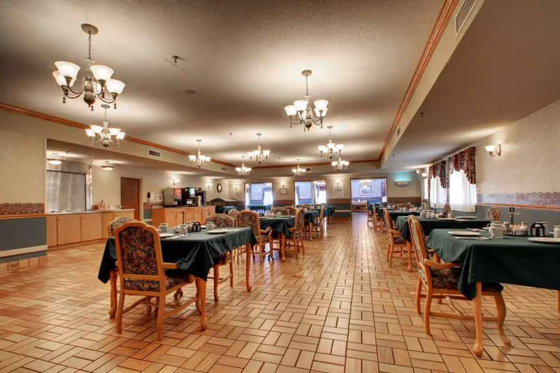 Large dining room with multiple tables covered in dark green tablecloths, wooden chairs, and chandeliers overhead.