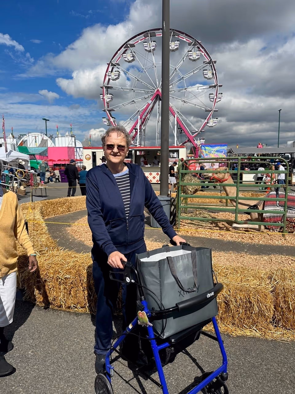An elderly woman using a blue walker with a gray bag attached, standing outdoors at a fair or carnival. Behind her is a large Ferris wheel, hay bales arranged as barriers, and a petting zoo area with animals in a fenced enclosure. The sky is partly cloudy with patches of blue.