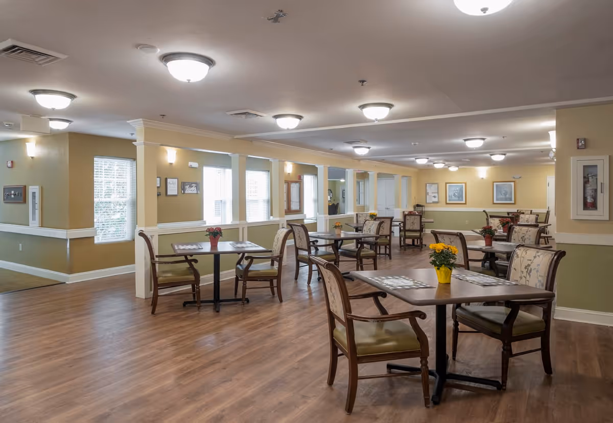 A spacious dining room in a senior living facility with multiple wooden tables and cushioned chairs arranged neatly. Each table has a small flower pot with colorful flowers. The room has warm yellow walls, large windows letting in natural light, and ceiling lights providing additional illumination. The floor is wooden, and there are framed pictures on the walls.