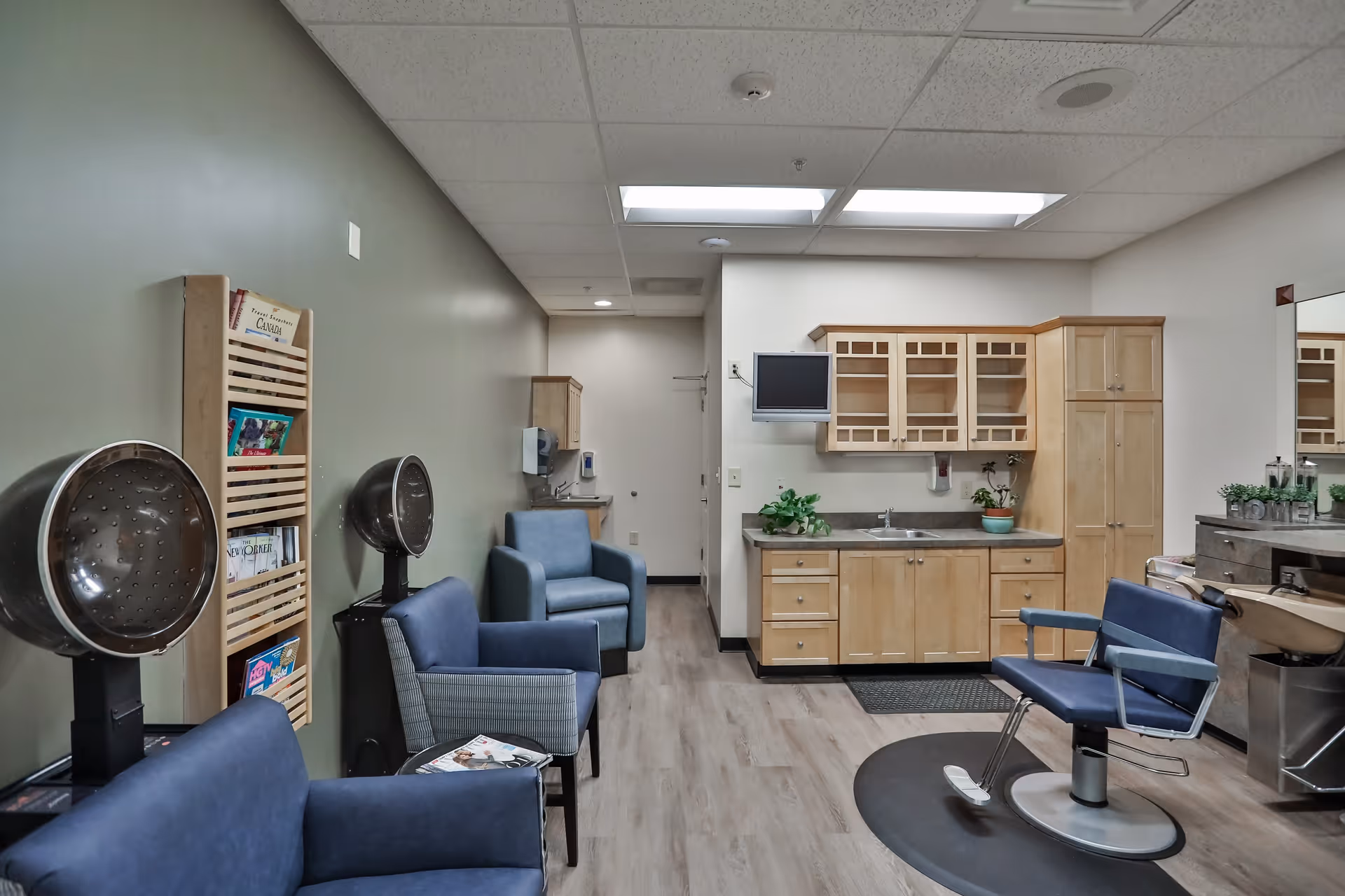 Interior of a senior living facility's salon area featuring blue salon chairs, hair dryers, a small kitchenette with wooden cabinets and a sink, a wall-mounted TV, and a magazine rack. The room has a light wood floor and neutral-colored walls.