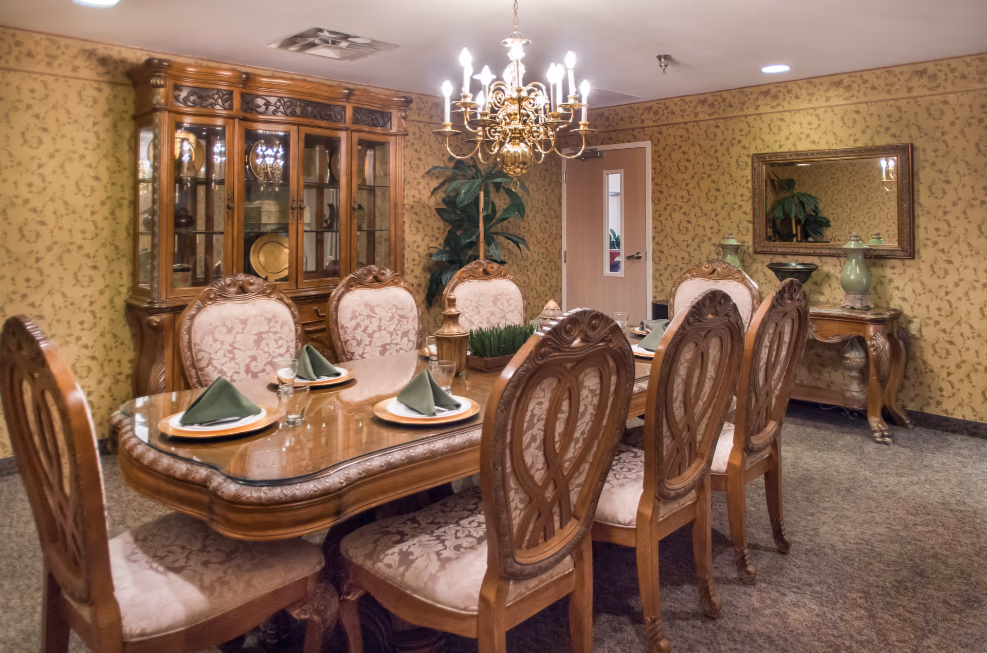 A formal dining room with an ornate wooden dining table set for six, featuring cushioned chairs with intricate wood carvings. The table is set with plates, glasses, and folded green napkins. A large wooden china cabinet with glass doors displays decorative items. The room has patterned wallpaper, a chandelier hanging above the table, a mirror on the wall, and a small wooden side table with decorative vases.