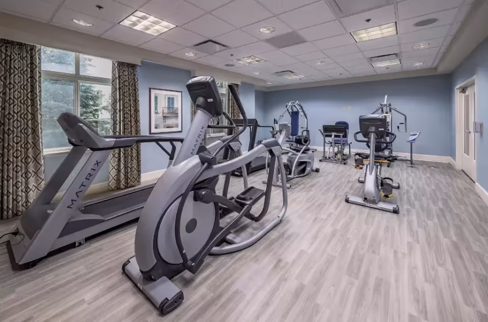 A fitness room in a senior living facility featuring various exercise equipment including treadmills, elliptical machines, and strength training machines. The room has light wood flooring, blue walls, large windows with patterned curtains, and a framed picture on the wall.