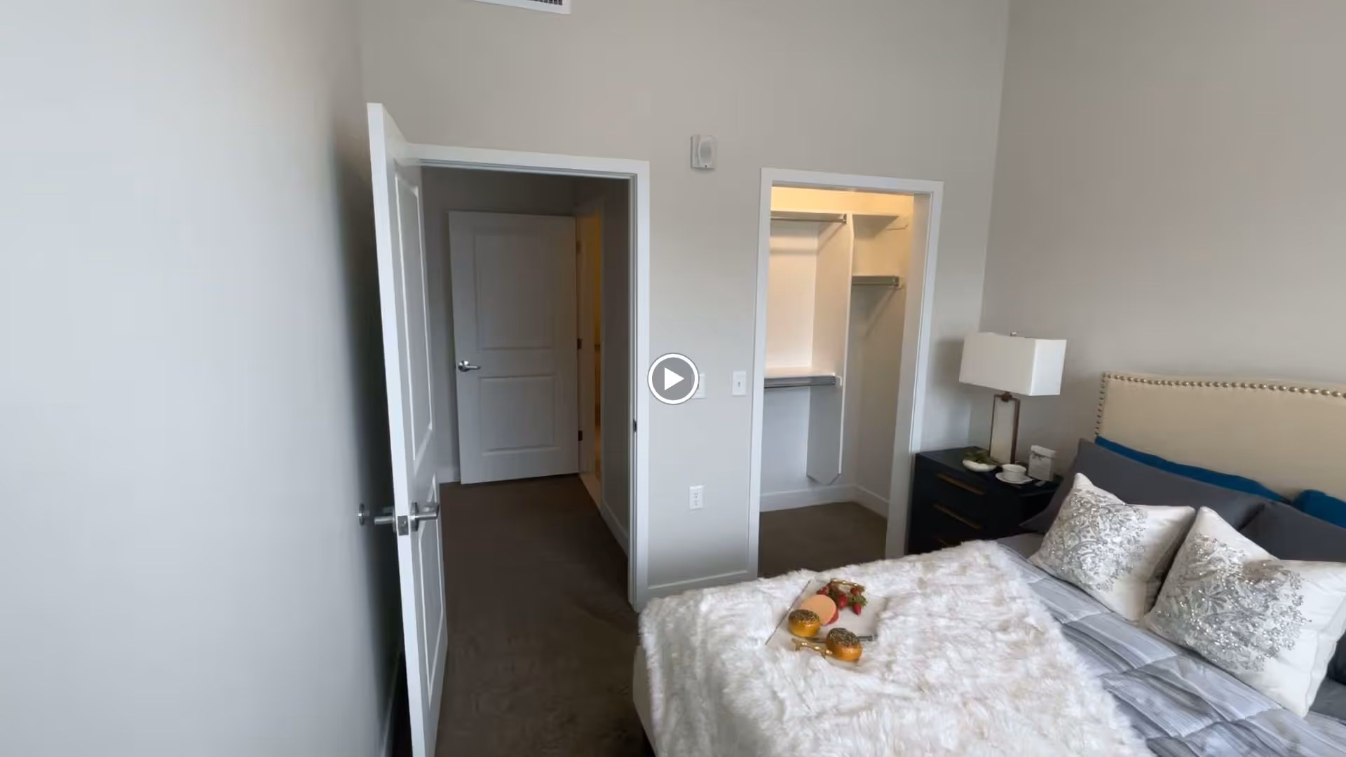 A bedroom in an assisted living facility featuring a neatly made bed with decorative pillows and a white fluffy blanket. There is a bedside table with a lamp and some items on it. The room has an open door leading to a hallway and another open door revealing a walk-in closet with shelves and hanging space.
