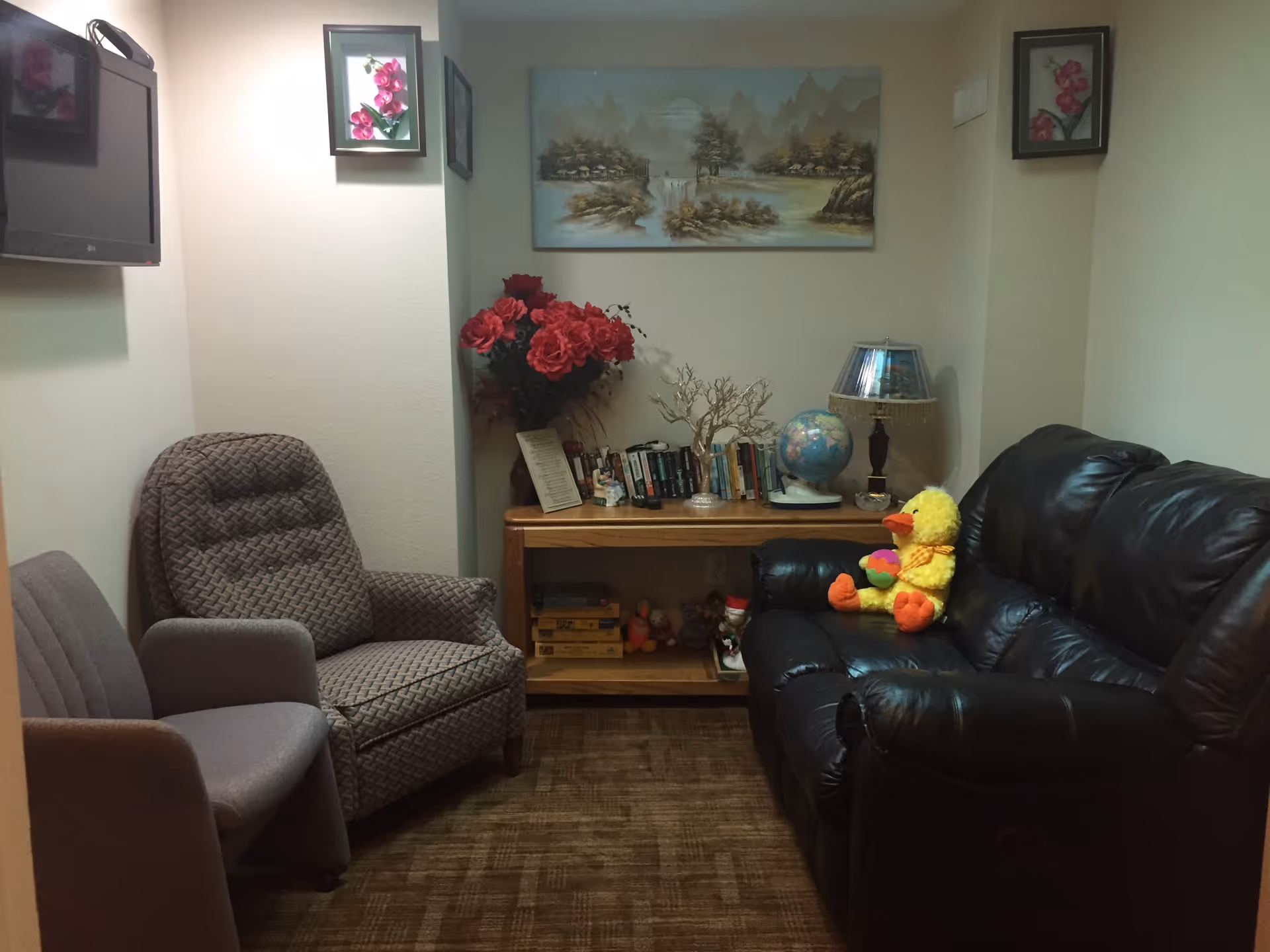 A small living room with a black leather couch on the right, a patterned armchair and a gray chair on the left. A wooden console table against the back wall holds books, a globe, a lamp, and a vase with red flowers. A stuffed yellow duck toy sits on the couch. The walls have framed floral and landscape paintings, and a flat-screen TV is mounted on the left wall.