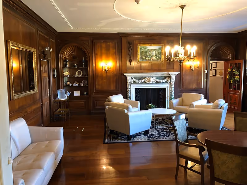 A cozy living room with wooden paneled walls, a white fireplace with decorative molding, and a chandelier hanging from the ceiling. The room features four white armchairs arranged around a small round table on a patterned rug. There is a white sofa on the left side and a wooden dining table with chairs on the right. A built-in wooden shelf with decorative plates and a water dispenser is visible in the background.