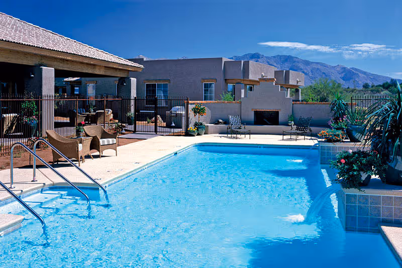 Outdoor swimming pool with clear blue water surrounded by a patio area with chairs and potted plants. In the background, there is a building with a covered seating area and a fireplace, with mountains visible under a blue sky.